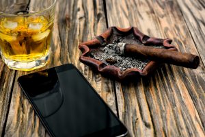 Saturated, contrasting photo of a glass of whiskey with ice, smoldering cigar on an ashtray and a smartphone on an old textured wooden table